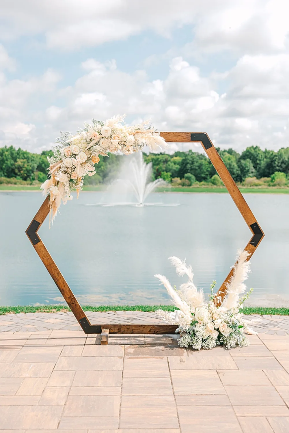 A hexagonal wooden wedding arch dressed in pampas grass and ivory roses, framing the lake fountain