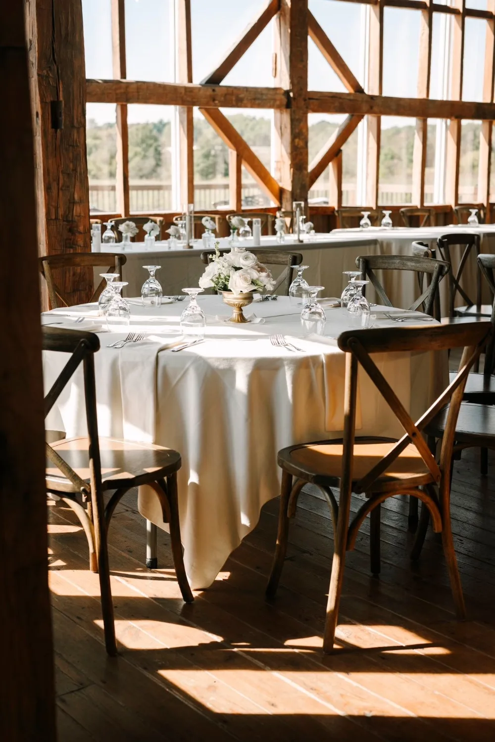 A sunlit reception table set with white linens, cross-back chairs and fresh florals